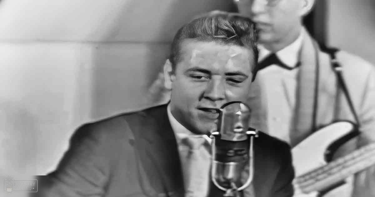 A young adult male singing into a microphone in a studio environment with an adult male in the background wearing a light-colored shirt or jacket.