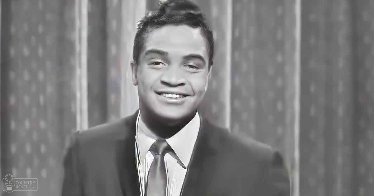 A young adult male wearing a dark suit jacket with a classic fit, a white dress shirt, and a neatly knotted tie stands against patterned curtains indoors; the scene appears to be daytime.