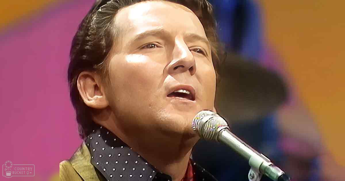 A middle-aged man wearing a dark-colored shirt with white polka dots and a gold or yellow jacket stands in front of a microphone at an indoor event, possibly performing or presenting.