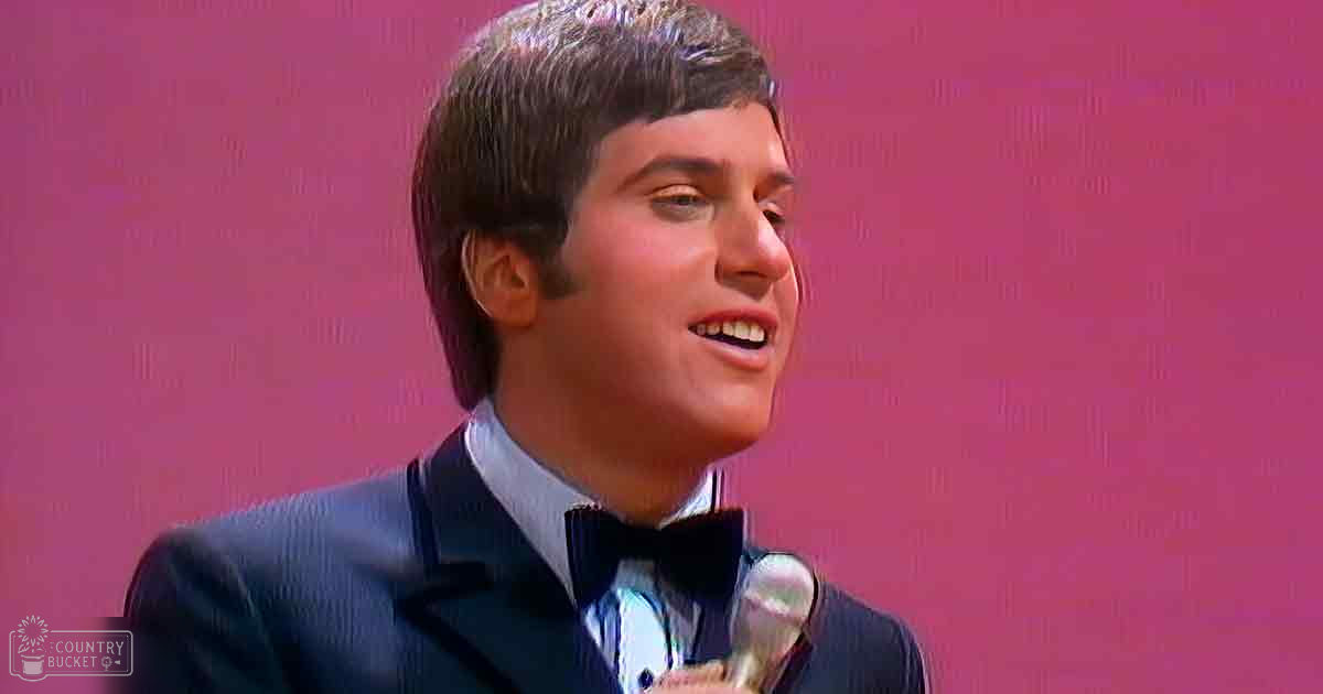 A young adult male in a tailored dark blue suit jacket and collared white shirt, set against a solid pink background with stage lighting suggesting an indoor event or performance space.