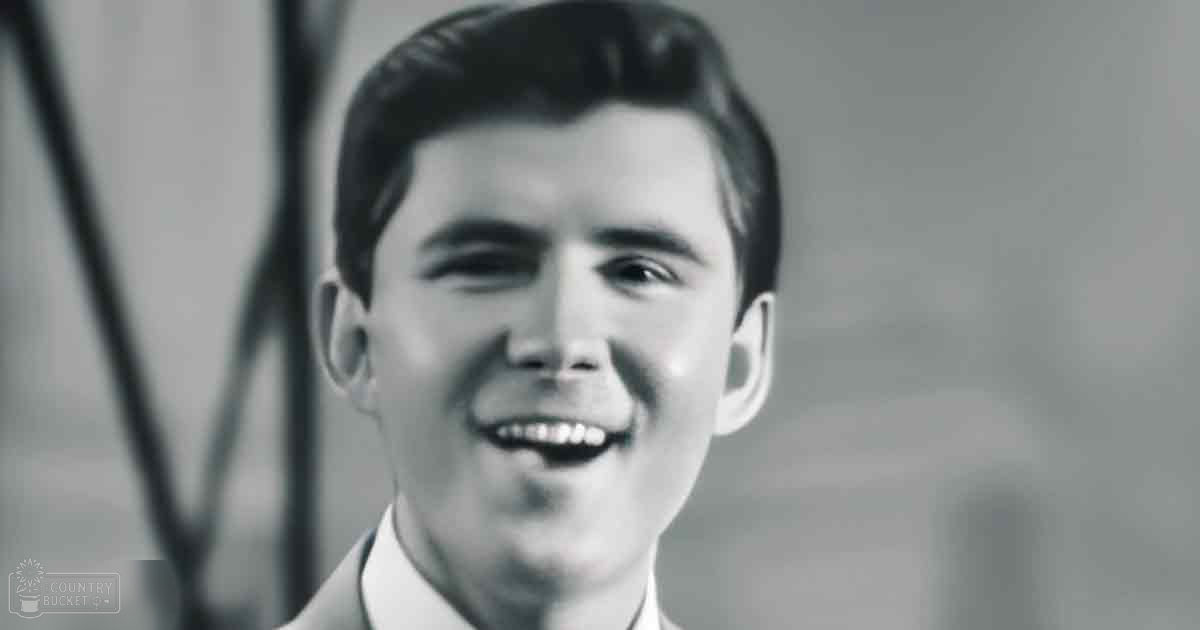A young adult male stands indoors, wearing a tailored suit jacket and white shirt in black-and-white photography.