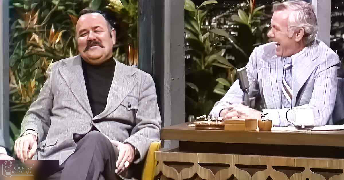 Two older adults in a talk show setting with large windows showing greenery outside, discussing on stage under natural light.