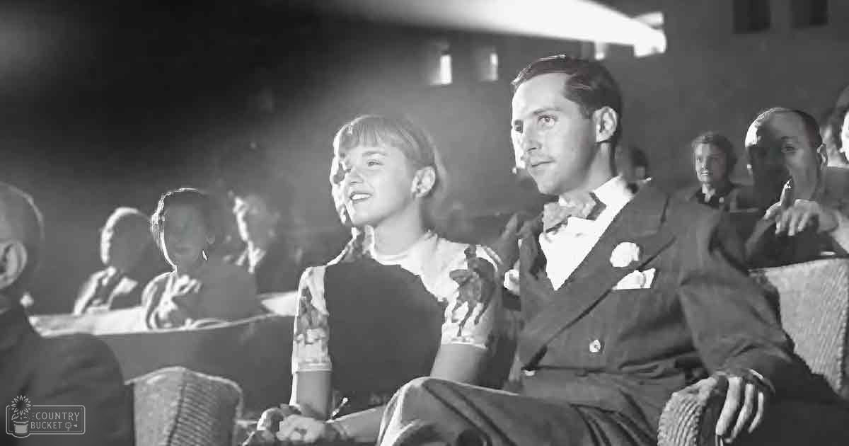 A mid-20th century couple in formal attire seated in an auditorium or theater with rows of seats, dimly lit and possibly evening time.