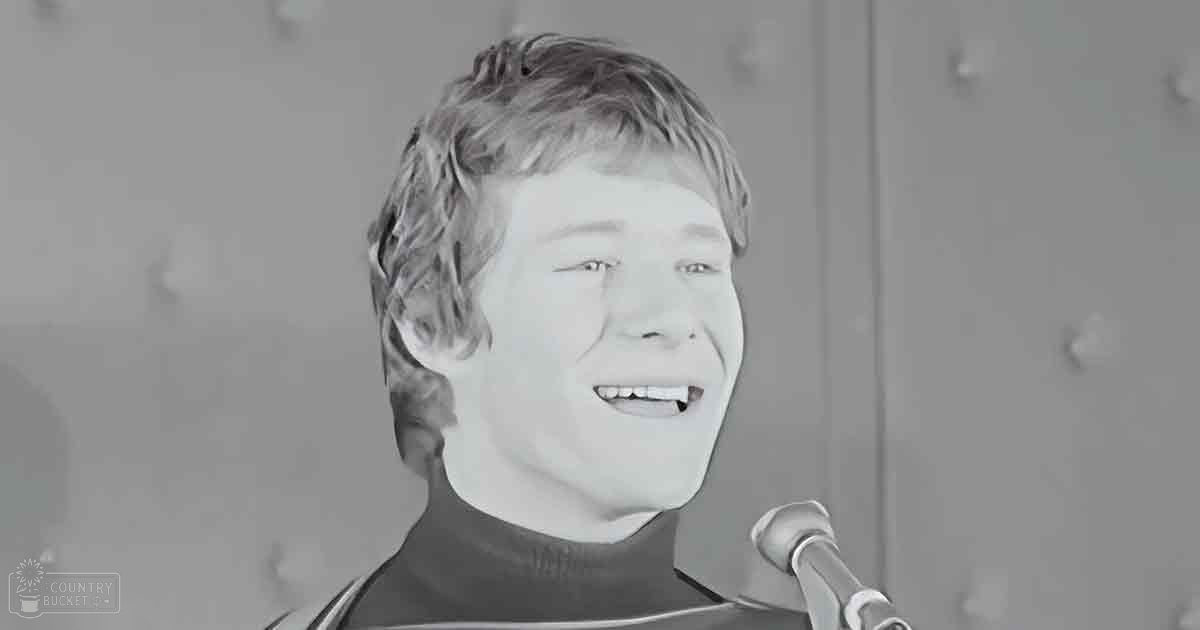 A young adult male wearing a dark turtleneck sweater stands indoors with a microphone in front of him; lockers and cabinets suggest a school setting, bright fluorescent lighting indicates daytime.