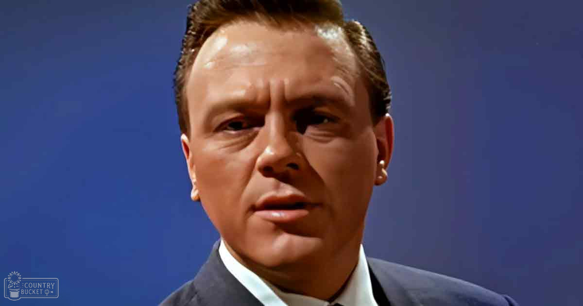 A middle-aged adult male in a dark blue suit jacket and white shirt against a plain blue backdrop, possibly a studio setting.