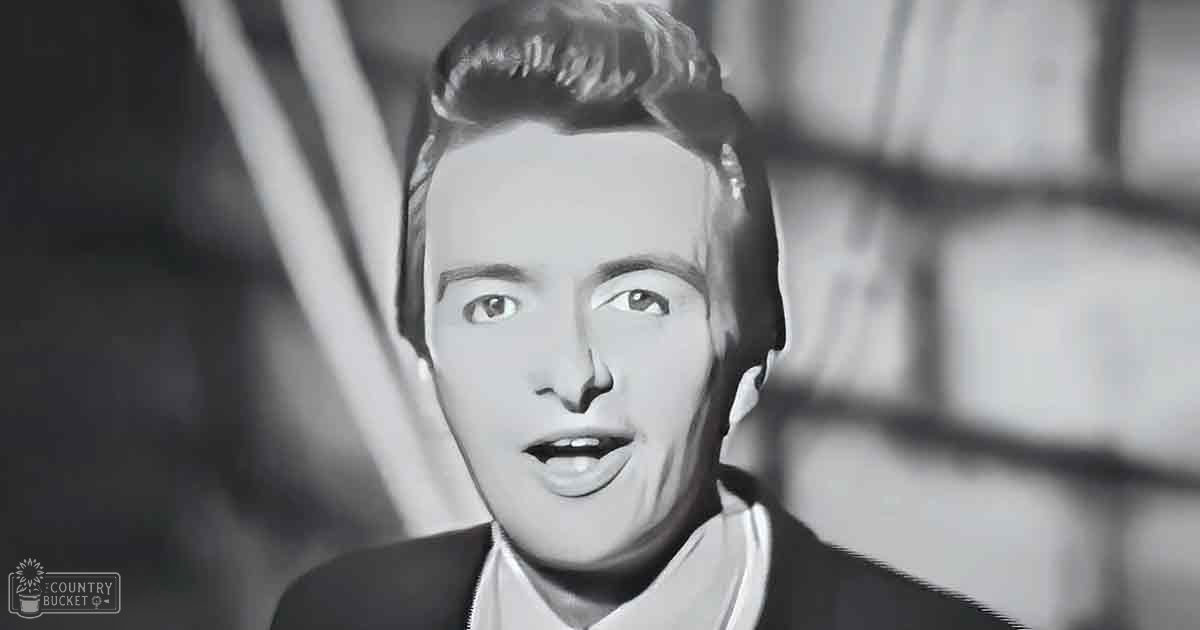 A young adult male wearing a dark suit jacket with a high-collared white shirt stands indoors against a blurred architectural background, possibly from an older film or photograph.