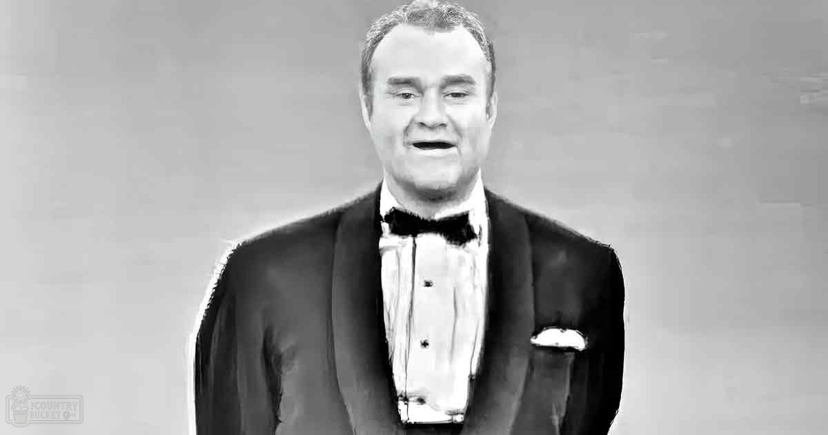 An adult male wearing a formal tuxedo with a bow tie, standing against a plain indoor background.
