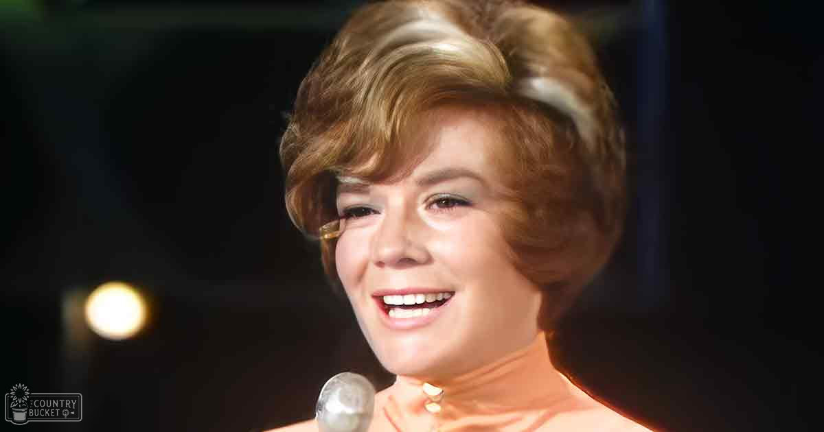 An adult female in a form-fitting, high-necked orange strapless dress or top stands indoors on what appears to be a stage with a microphone and lighting visible. She wears a small pendant necklace.