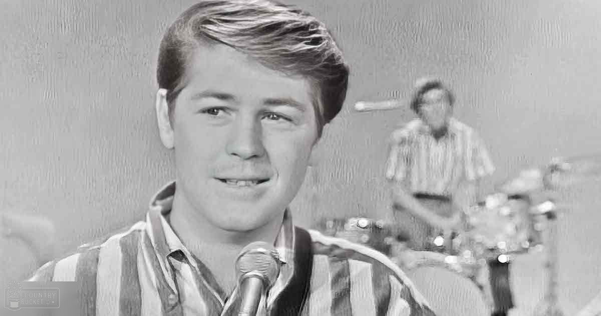 A young adult male wearing a striped shirt performs on stage with an adult male drummer behind him, both set against neutral-colored indoor walls in a well-lit studio environment.