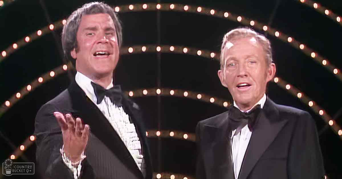 Two adult men in a formal setting, possibly on stage or at an event venue, wearing black tuxedos with white shirts and black bow ties.