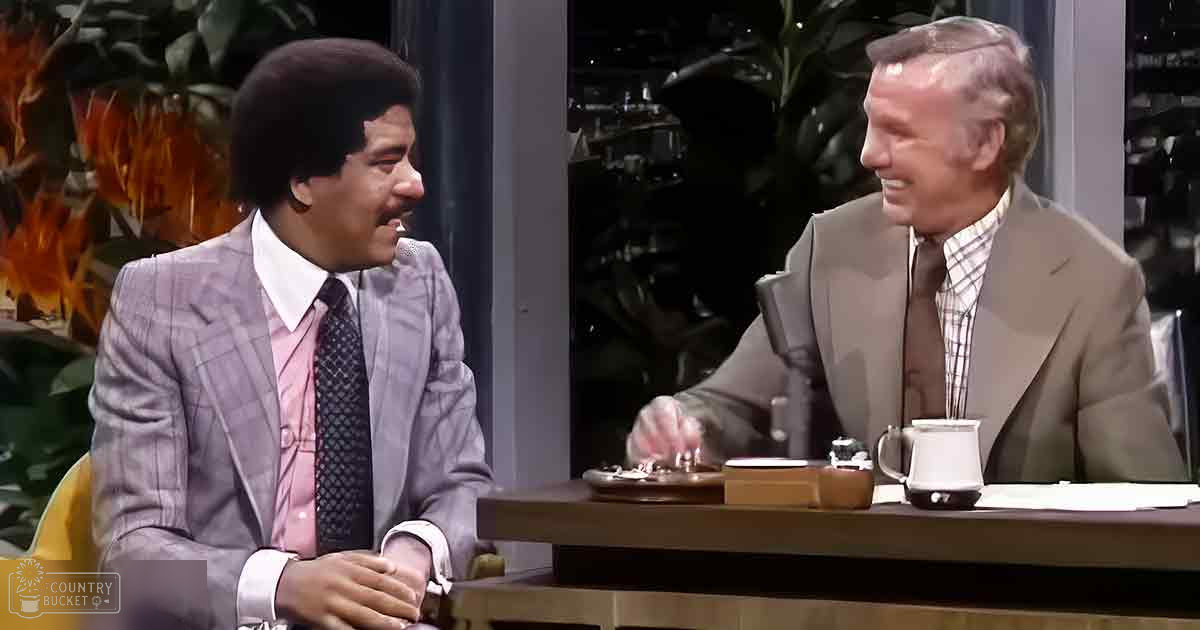 A talk show host and an older adult male in a studio setting with professional equipment and plants. The host wears a light gray suit, while the other man is dressed in a beige jacket over a checkered shirt.