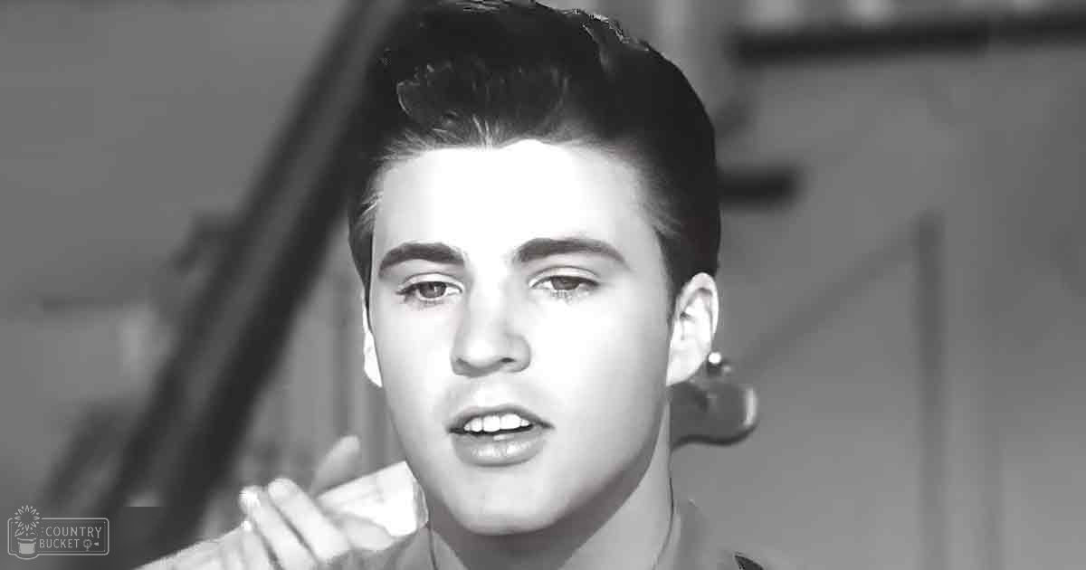 A young adult male with styled hair, wearing a mid-20th-century collared shirt indoors.