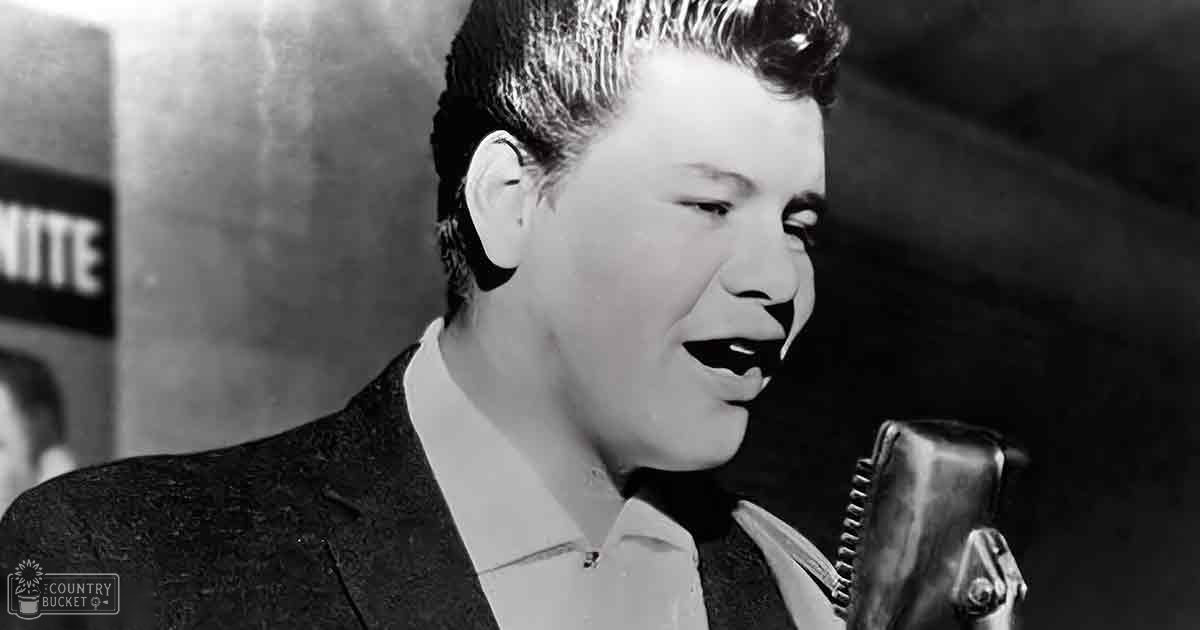 A young adult male singer in a dark suit jacket with a white shirt, earpiece visible, singing into a microphone against an indoor backdrop.