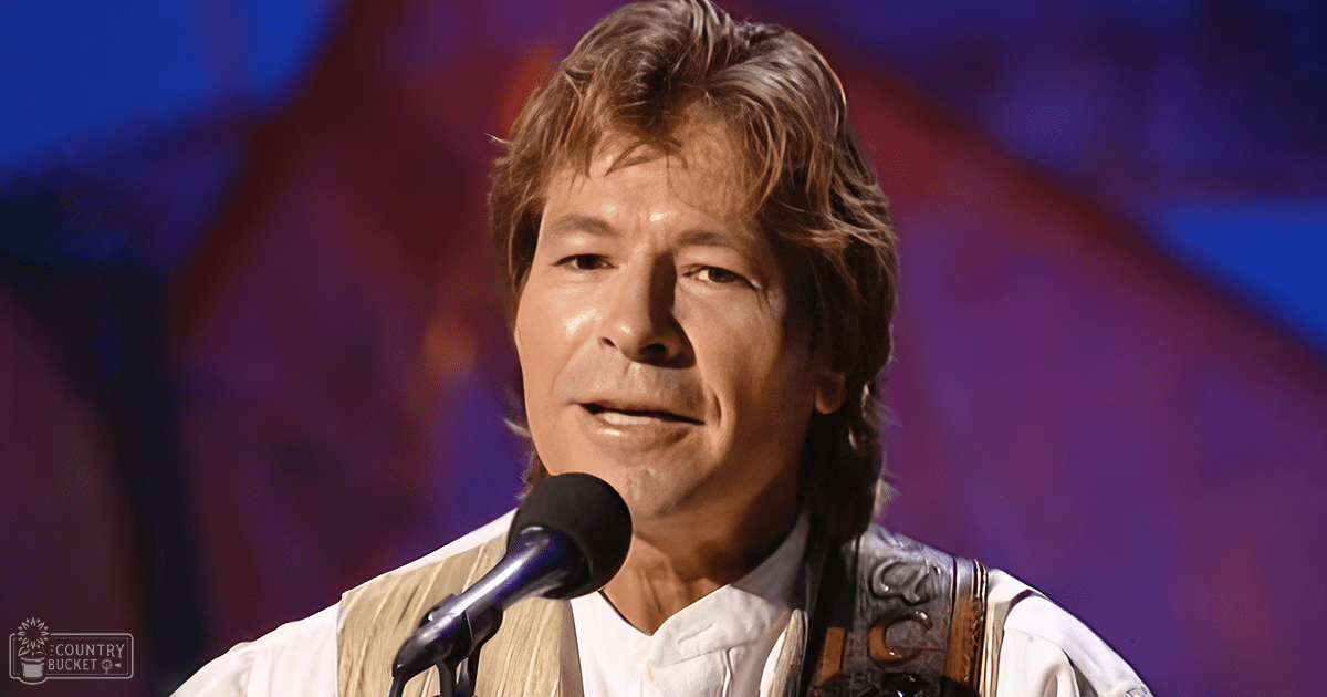 A middle-aged adult male in a modern casual white shirt with long sleeves, standing on stage under artificial blue and purple lighting during an indoor event.