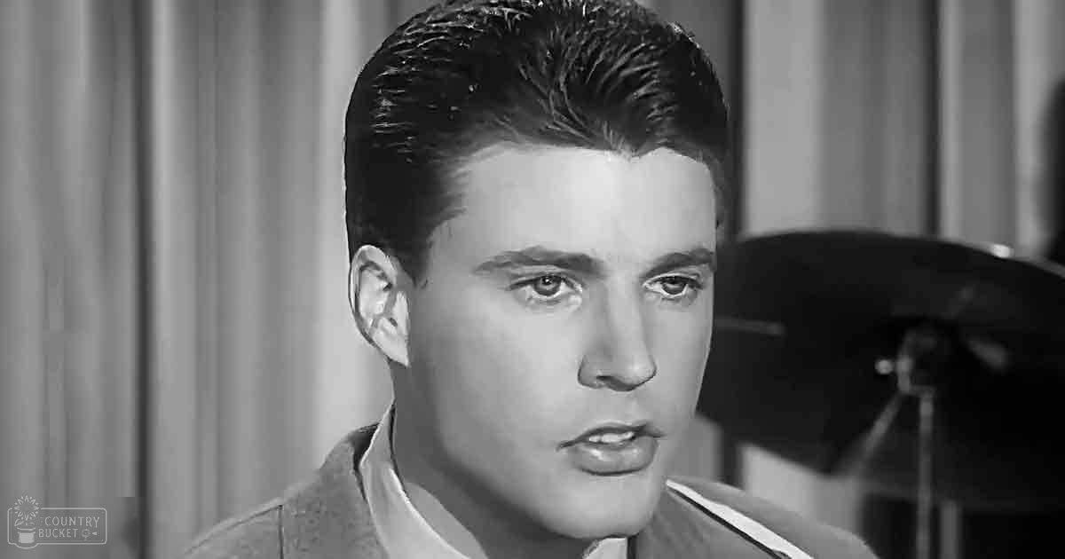 A young adult male dressed formally in a mid-20th century style collared shirt and textured jacket, with a curtain backdrop suggesting an indoor setting possibly on a stage.