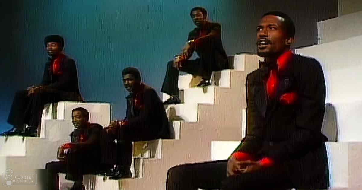 An adult male singer seated in front, wearing dark suit attire with red shirt, indoors on stage-like setting with controlled lighting and muted tones.
