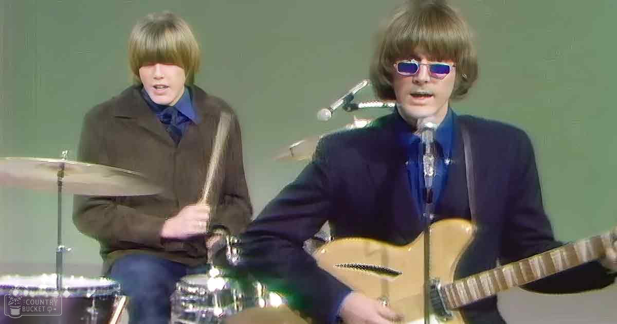 A music performance scene with two young adult males, one in a brown jacket and the other in a dark blue suit, with a drum set and microphone stand visible against a plain backdrop.