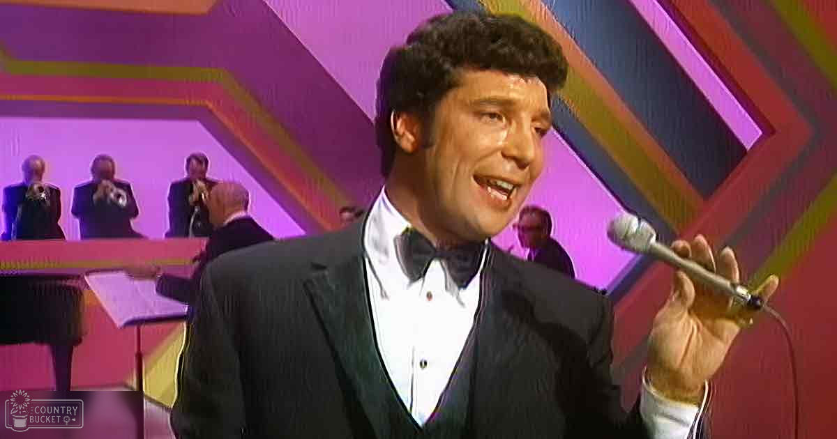 A middle-aged male in a black tuxedo stands at a piano, holding a microphone under controlled indoor lighting, possibly during a performance or rehearsal.