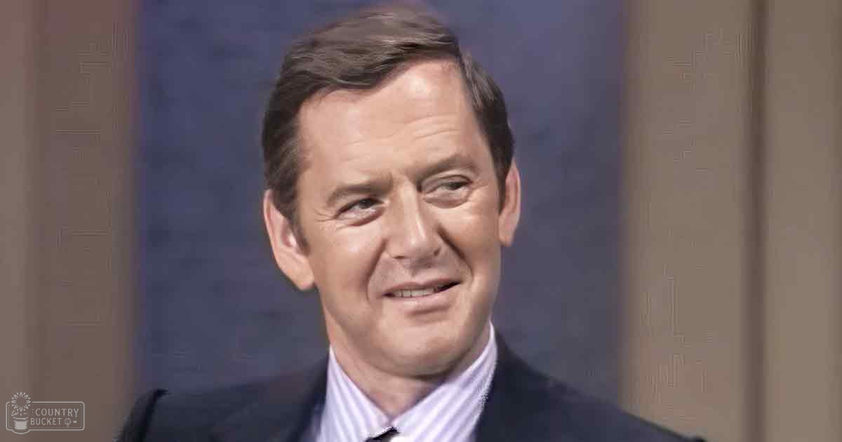 Middle-aged man wearing dark suit jacket, white collared shirt with light stripes, and tie, standing indoors against a plain wall background.