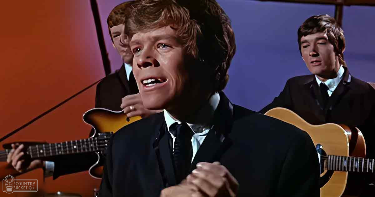Young adult male in black suit jacket and white shirt, performing with other similarly dressed individuals against an indoor stage backdrop.