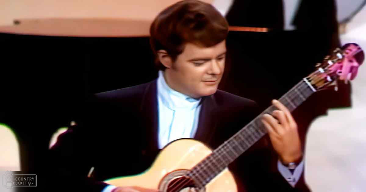 A male adult in a dark suit jacket with white shirt and light tie plays an acoustic guitar against a backdrop of dark curtains, likely at a concert or performance venue.