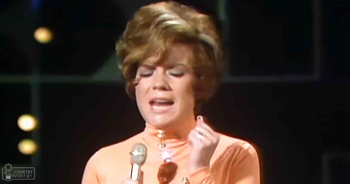 A female adult singer in a peach-colored, mid-20th-century-style top performs on an indoor stage with blurred stage lights and microphone setup.