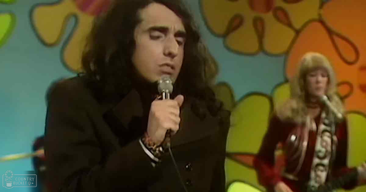 A male performer in a black suit jacket over a white shirt holding a microphone, standing against a vibrant floral backdrop with psychedelic patterns at what appears to be an indoor concert or performance venue from the late 1960s-early 1970s.