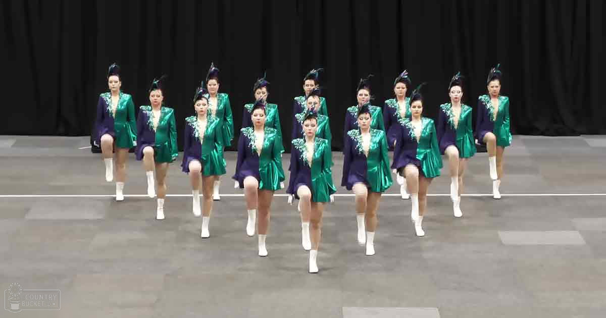 A group performance in an indoor arena or gymnasium with young adult female dancers wearing form-fitting, green and purple modern dance costumes and white knee-high boots.