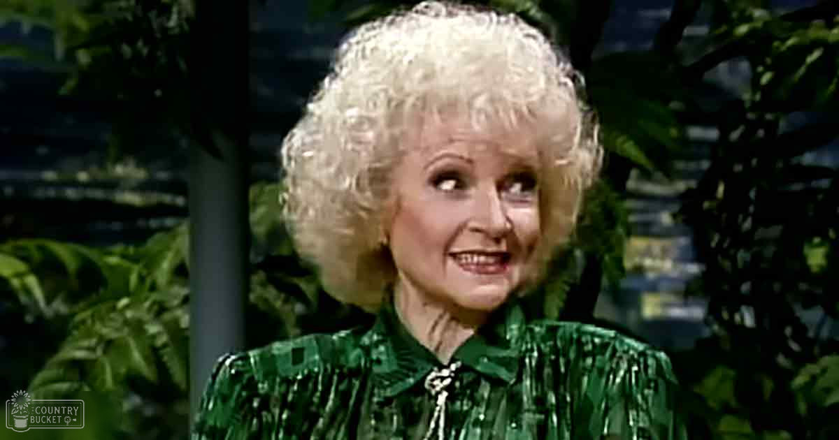 An older adult female in a garden setting, wearing a green patterned blouse with ruffled details around the collar and sleeves.