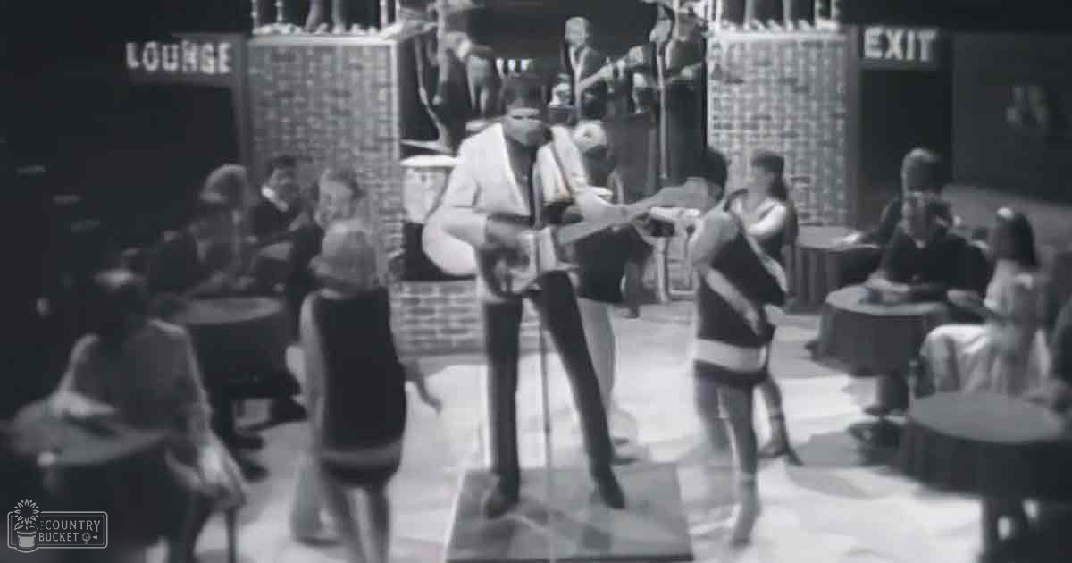 Adult male performer in a lounge setting playing guitar with an audience seated around him; vintage attire suggests mid-20th century ambiance.