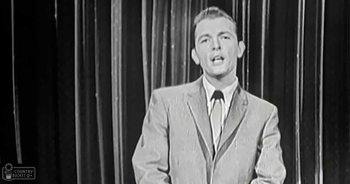 Young adult male wearing a light-colored suit jacket with notched lapels, standing indoors against vertical curtains.