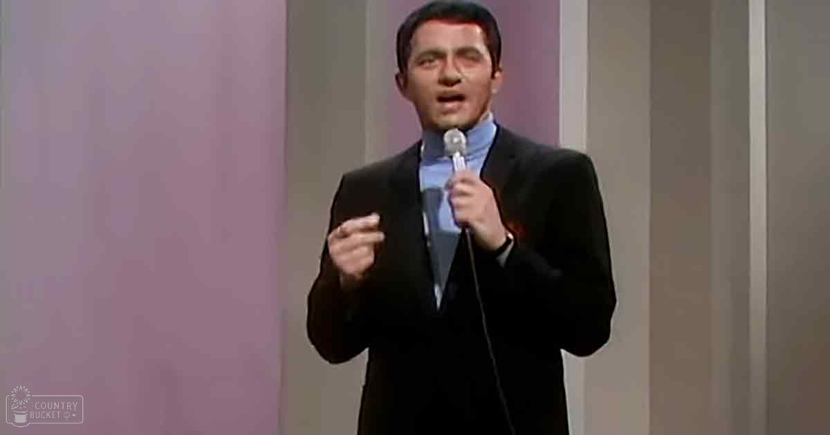 An adult male performer in a dark suit jacket, light blue collared shirt, holding a microphone against soft indoor lighting with stage panels behind him.