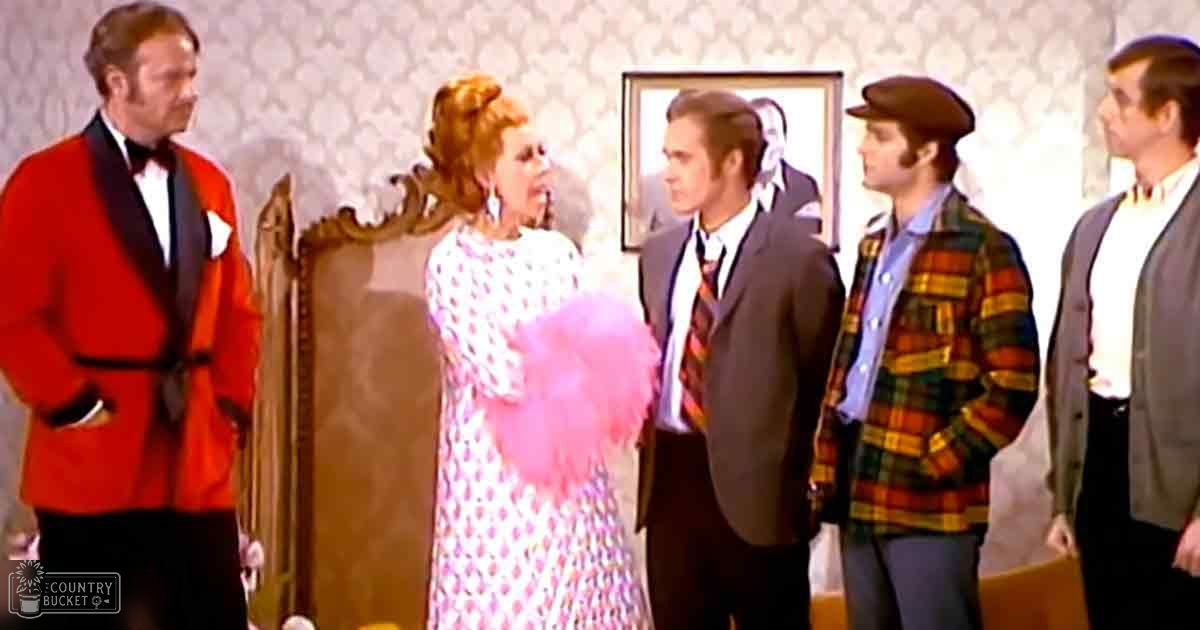 A formal indoor gathering with adults in suits and dresses; red blazer, patterned pink dress, dark suit jacket, plaid jacket, gray suit jacket;