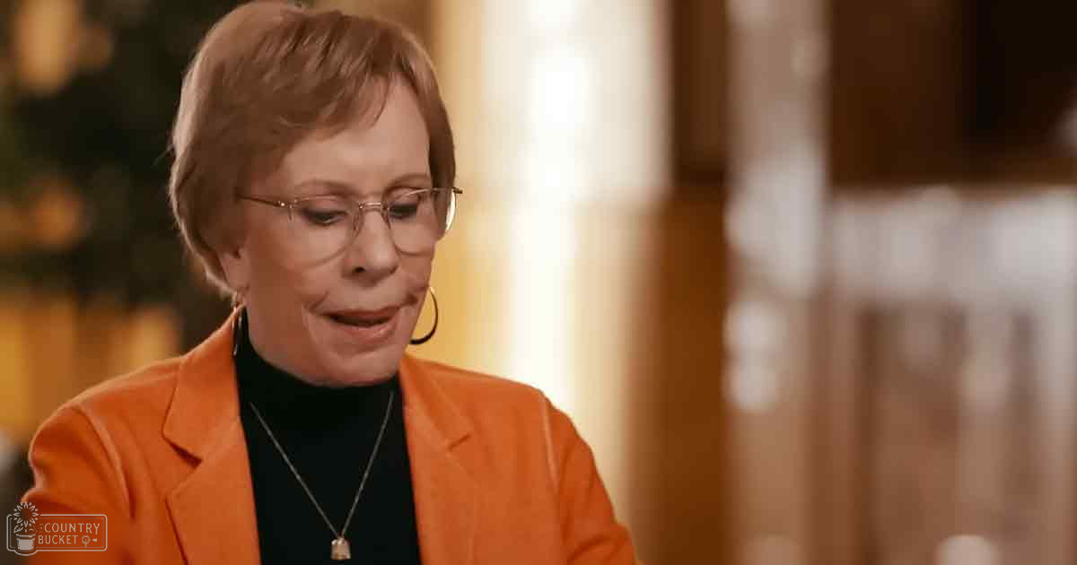 A middle-aged to older adult female in a home setting, wearing an orange blazer and black turtleneck sweater. The scene is illuminated by natural light with beige curtains visible in the background.
