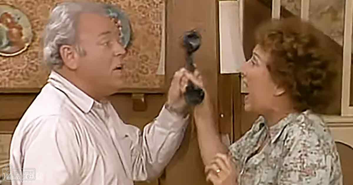 A middle-aged man holds a small figurine near his face as an older woman looks on, set against warm-toned wall decorations and wooden furniture in what appears to be a home interior.