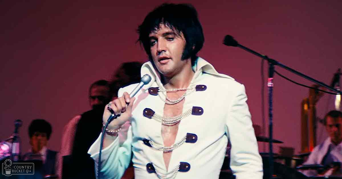 A mid-20th century performer in a white suit with purple buttons at a concert venue, surrounded by other adults in formal attire; stage lighting and simple red/maroon backdrop suggest an indoor setting.