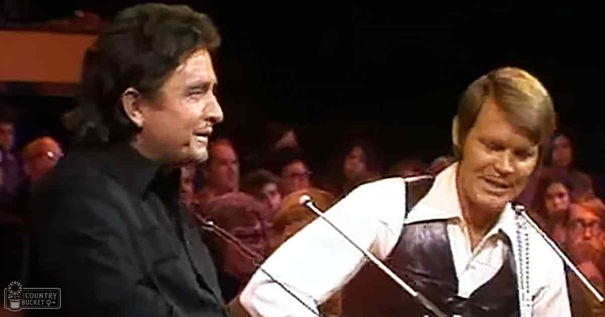 Musical performance by two adults, one wearing a fitted black shirt and the other an older adult male in a white button-down under a brown leather vest, with a microphone stand indicating singing or speaking together on stage.