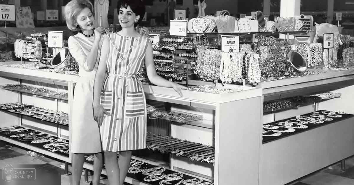 Two young adult females in a mid-20th-century jewelry store, wearing fitted dresses with flared skirts; shelves of necklaces and price tags visible.