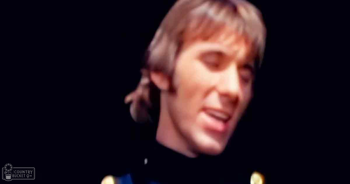 A young adult male stands indoors wearing a dark-colored garment with visible buttons from the 1960s or early 1970s, set against a dark, low-lit background.