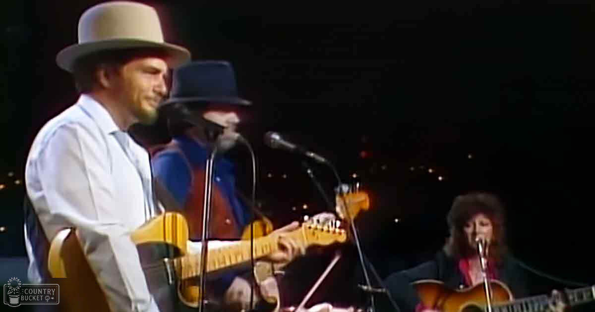 A live musician performs on stage under artificial lighting; an adult male singer wears a light-colored shirt and wide-brimmed hat.