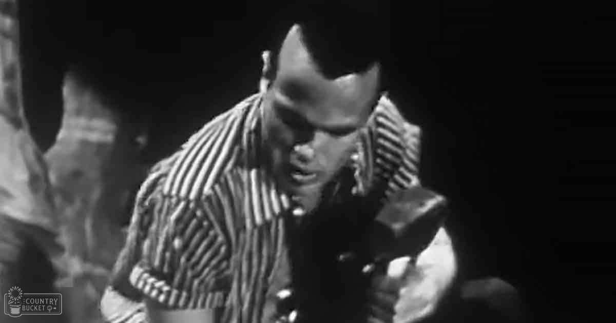 An adult male wearing a striped shirt indoors with subdued lighting; another person partially visible on the left side in a robe or jacket.