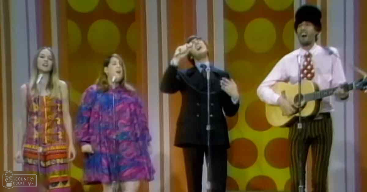 A musical performance scene with performers in late 1960s or early 1970s attire, set against a backdrop of large yellow circles on an orange and white background.