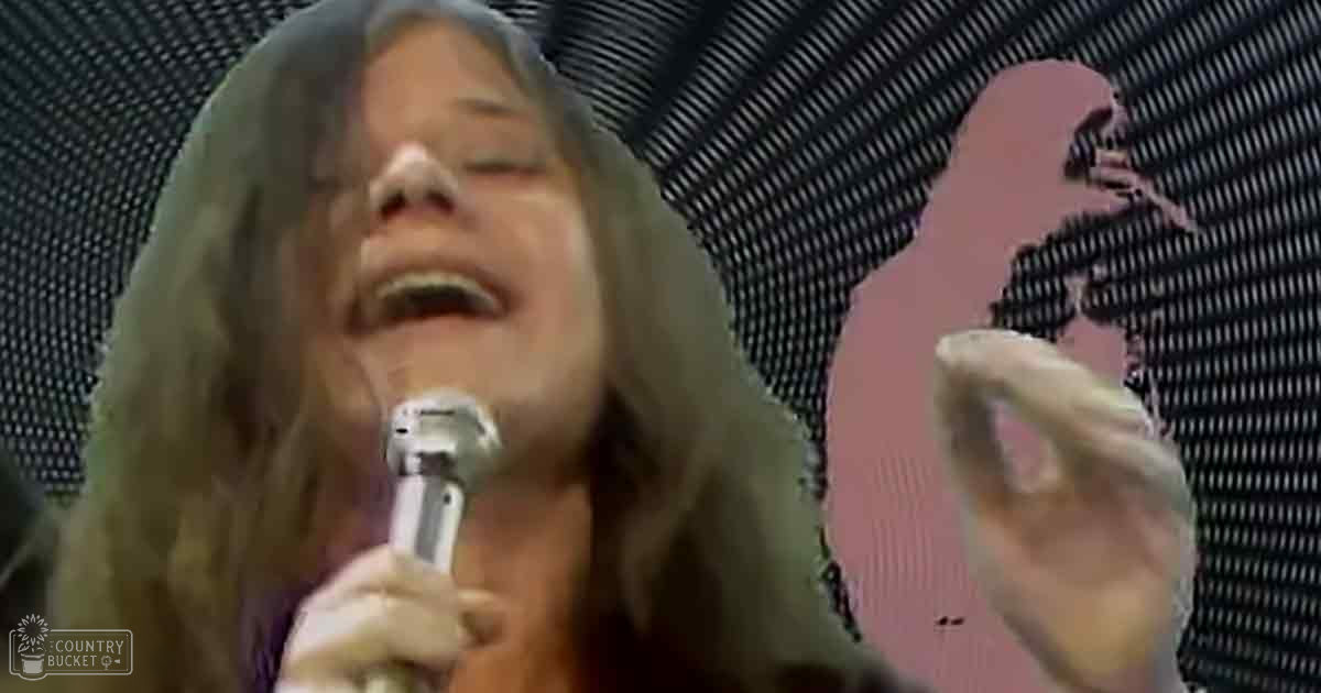 An adult female performer singing into a microphone at what appears to be a concert or event; wearing a low-cut, patterned blouse from the 1960s/early 70s with semi-transparent fabric and floral elements.