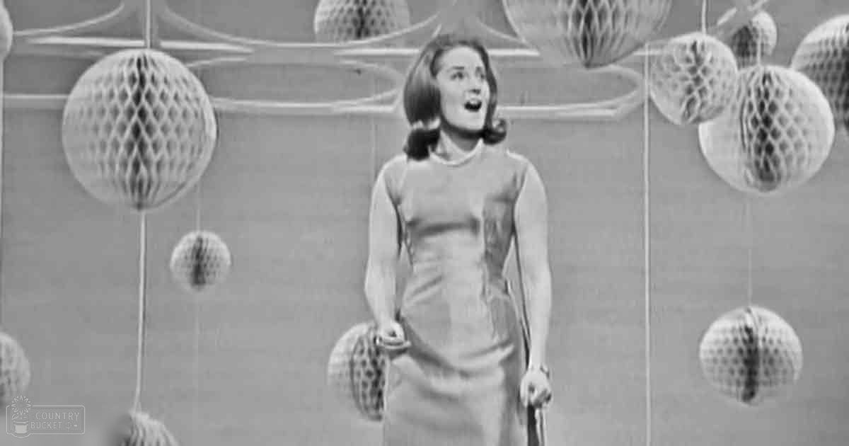 A young adult female in a vintage-style dress holds a clutch purse indoors with hanging decorative items suggesting a festive atmosphere.