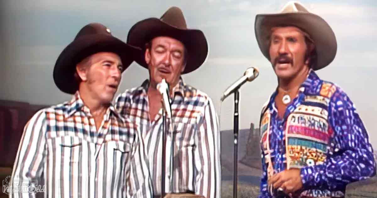 An outdoor musical performance with three adult males in plaid shirts and cowboy hats under an overcast sky.