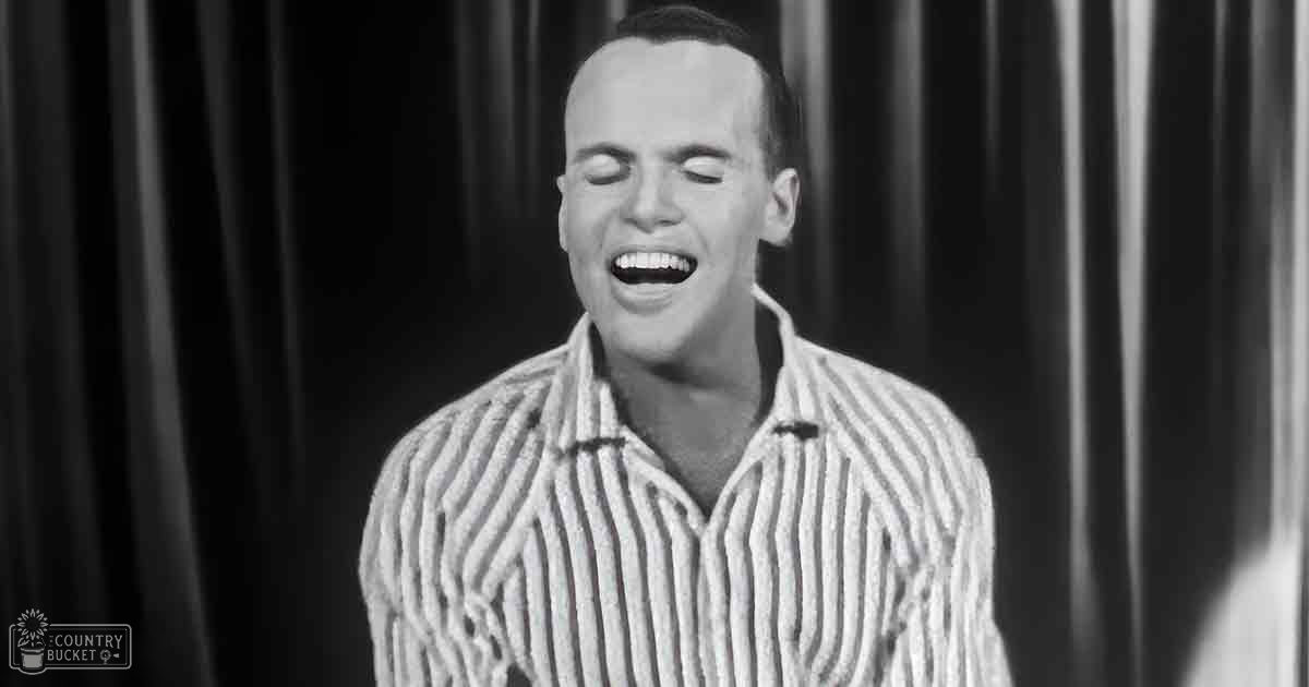 A young adult male wearing a relaxed, casual striped shirt with vertical lines in mid-20th century style stands indoors against dark curtains.