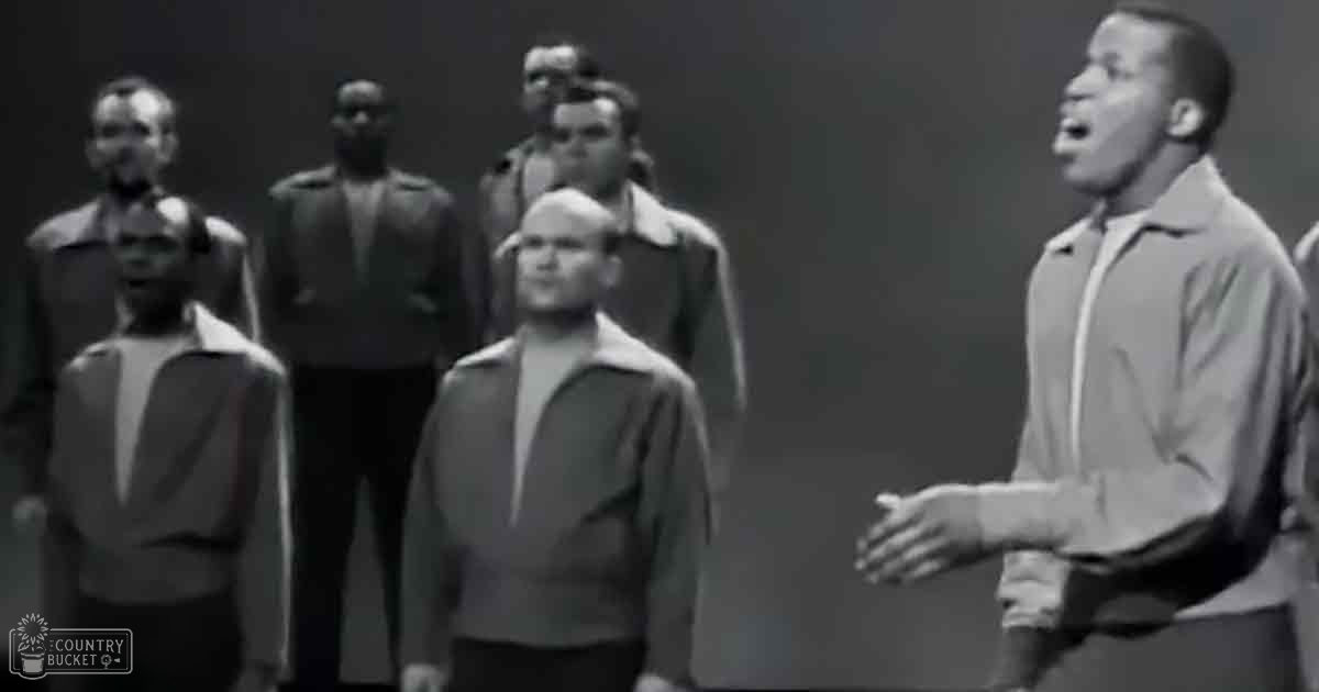 A group of men singing in a studio setting with plain dark walls, even lighting suggesting daytime.