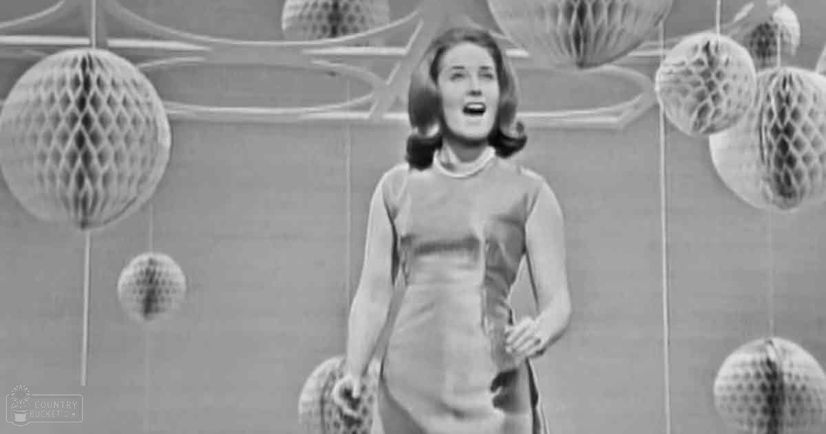 A young adult female wearing a form-fitting, long-sleeved dress stands indoors with decorative spherical items hanging from strings against a plain wall. The setting appears to be an indoor space designed for performances or presentations.