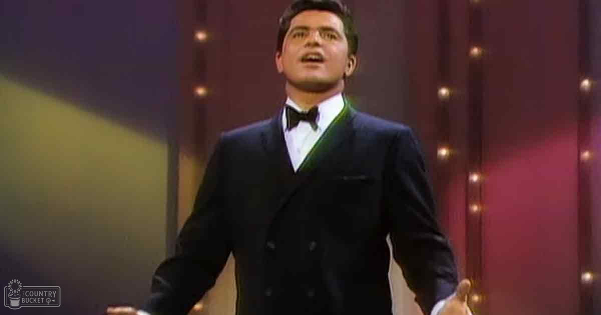 An adult male singer wearing a dark double-breasted suit jacket with a white shirt and black bow tie, performing indoors under artificial stage lighting.