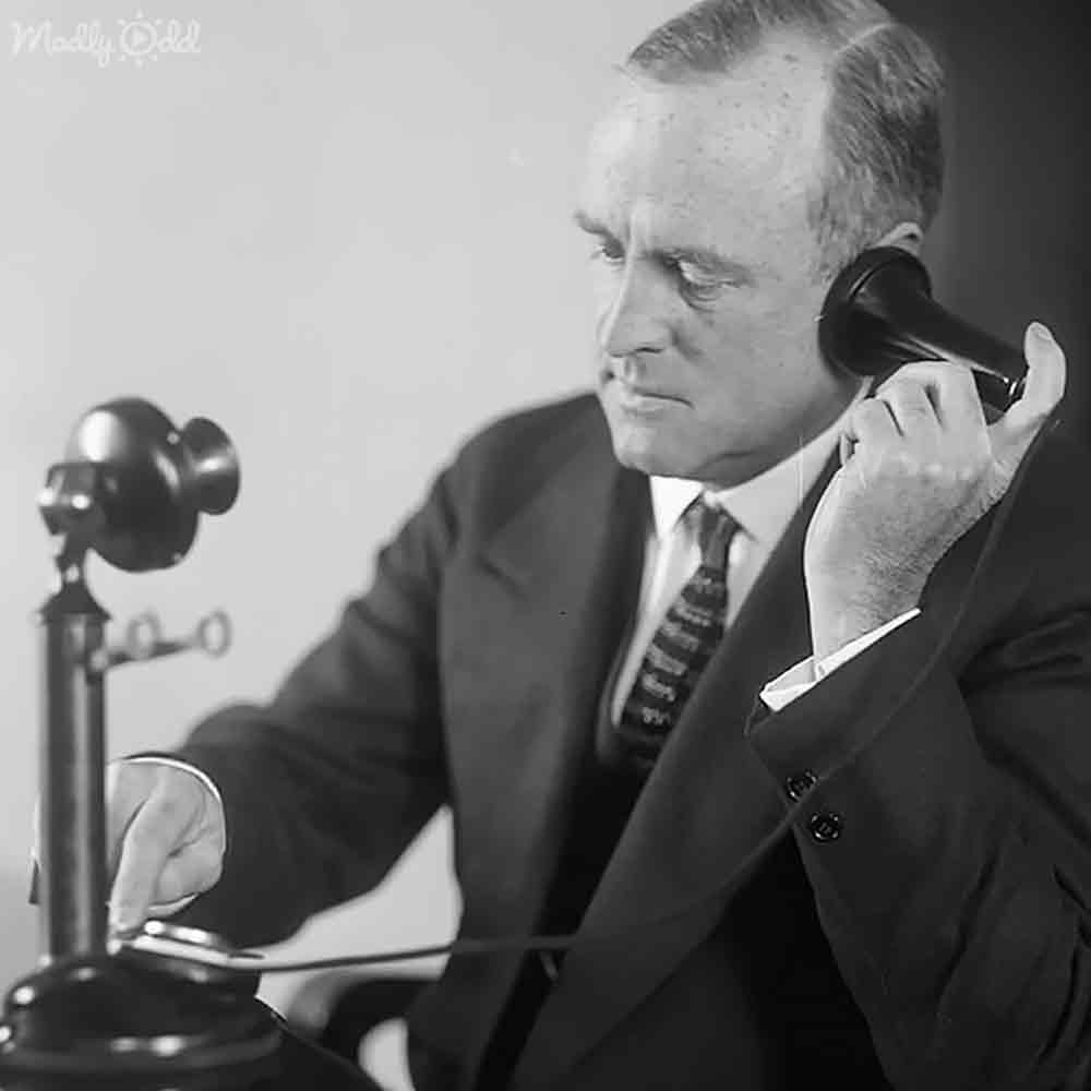 An older adult male wearing a dark suit jacket, white dress shirt, and patterned tie sits at an old-fashioned telephone desk under artificial light.